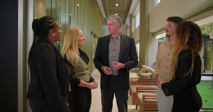 Corporate manager speaking to diverse team in modern office hallway, group of professionals engaged in workplace conversation during informal meeting