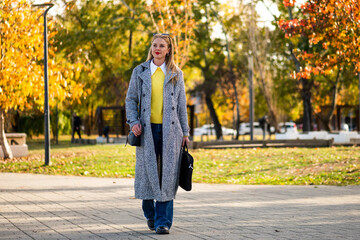 Confident and urban businesswoman in coat walking through sunny autumn park, wearing sunglasses and carrying laptop bag.