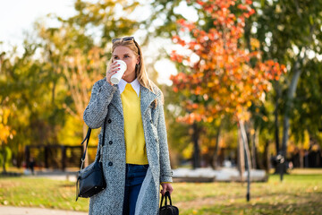 Modern businesswoman in autumn coat holding laptop bag and drinking coffee while walking through city park during sunny day.