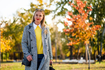 Modern businesswoman wearing sunglasses and stylish autumn coat while walking with a laptop bag through the city park during sunny day.