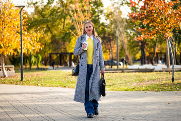 Urban businesswoman in autumn coat holding laptop bag and drinking coffee while walking through city park during sunny day.