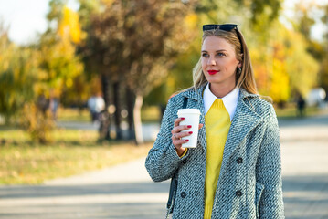 Portrait of modern businesswoman with sunglasses in stylish autumn coat drinking coffee while standing in the city park during sunny day.
