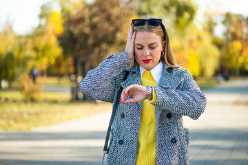 Stressed and worried businesswoman in autumn coat checking the time on her wristwatch while standing in the city park.