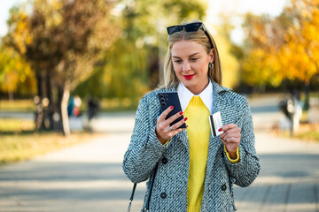 Modern businesswoman in coat using smartphone and credit card while standing in the city park during sunny autumn day. 