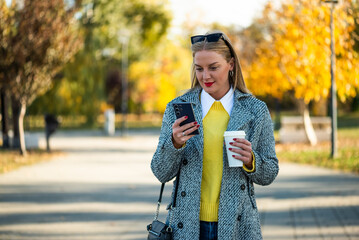 Portrait of urban businesswoman with sunglasses in modern autumn coat using smartphone and drinking coffee while standing in the city park during sunny day.