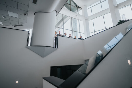 A diverse group of colleagues stands on a bright balcony in a modern office, overlooking a glass atrium and escalator. The scene conveys collaboration, curiosity, and professional energy. - Powered by Adobe