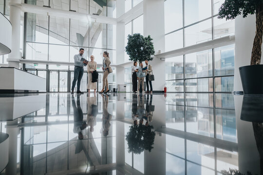 A diverse group of people gathers in a bright, modern office lobby to discuss ideas and collaborate. Glass walls, reflective floors, and indoor plants create a welcoming, professional atmosphere.