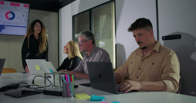 Corporate team works attentively during office presentation, with everyone focused on laptops and screen displaying data charts and business analytics - Powered by Adobe