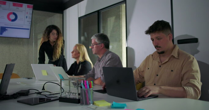 Corporate team works attentively during office presentation, with everyone focused on laptops and screen displaying data charts and business analytics