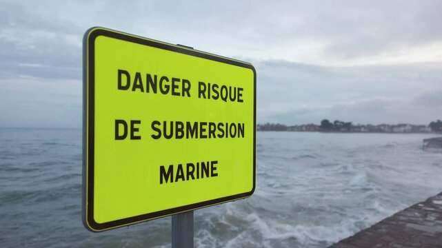 Fluorescent yellow coastal warning sign for marine submersion risk in Brittany, France, with rough Atlantic Ocean waves in the background, horizontal shot.