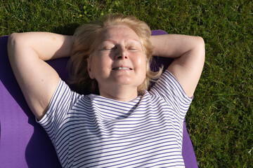 Senior woman lying on a yoga mat outdoors, relaxing on the grass with eyes closed and enjoying the warm sunlight. Peaceful moment of rest, wellness, and outdoor lifestyle.