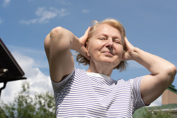 Senior woman standing outdoors with hands near her head, smiling softly and enjoying the warm sunlight against a clear blue sky. Concept of wellbeing, positive aging, and relaxed outdoor lifestyle.