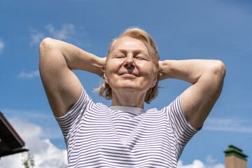 Senior woman enjoying sunlight outdoors with closed eyes, holding her hands near her head and smiling peacefully against a clear blue sky. Concept of wellbeing, relaxation, and positive aging.