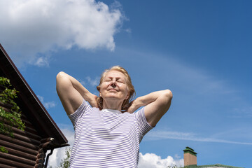 Senior woman standing outdoors with hands near her head, smiling softly and enjoying the warm sunlight against a clear blue sky. Concept of wellbeing, positive aging, and relaxed outdoor lifestyle.