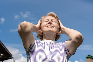 Senior woman enjoying sunlight outdoors with closed eyes, holding her hands near her head and smiling peacefully against a clear blue sky. Concept of wellbeing, relaxation, and positive aging.