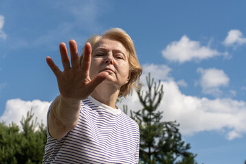 Elderly woman extending hand toward camera on a sunny day with blue sky and trees in background, stop gesture