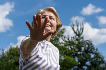Senior woman reaching her hand forward toward the camera outdoors, standing under a blue sky with clouds. Strong expressive gesture conveying stop, attention, or protection.