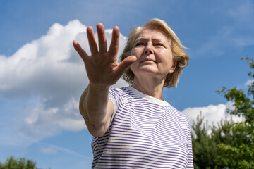 Senior woman reaching her hand forward toward the camera outdoors, standing under a blue sky with clouds. Strong expressive gesture conveying stop, attention, or protection.