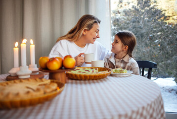 Woman and girl enjoying cozy winter breakfast at a table with pastries, apples, and candles, creating a warm family atmosphere with soft natural light
