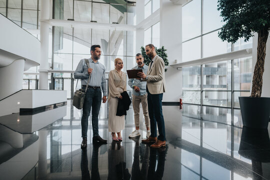 A diverse group of people stands in a bright, glass-walled lobby examining a tablet together, signaling collaboration and teamwork in a contemporary corporate setting. - Powered by Adobe