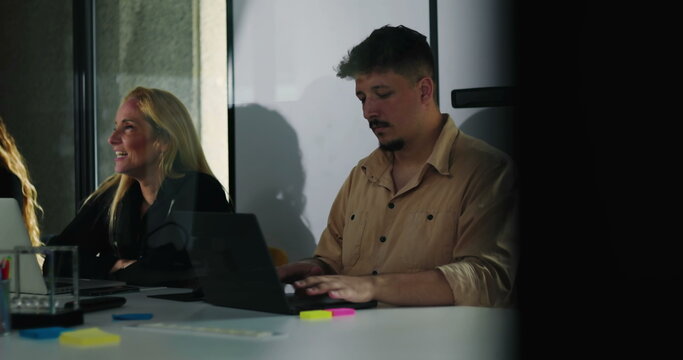 Coworkers quietly concentrate on laptop tasks during team meeting in bright office environment with natural daylight and modern collaborative setup