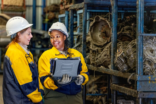 Two female engineers in safety gear inspecting car parts inventory. workers technical walking old dirty car industry checking used auto parts by tablet industrial automotive parts warehouse Recycling - Powered by Adobe