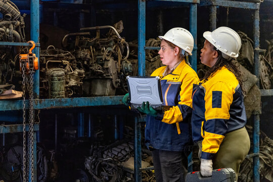 Two female engineers in safety gear inspecting car parts inventory. workers technical walking old dirty car industry checking used auto parts by tablet industrial automotive parts warehouse Recycling