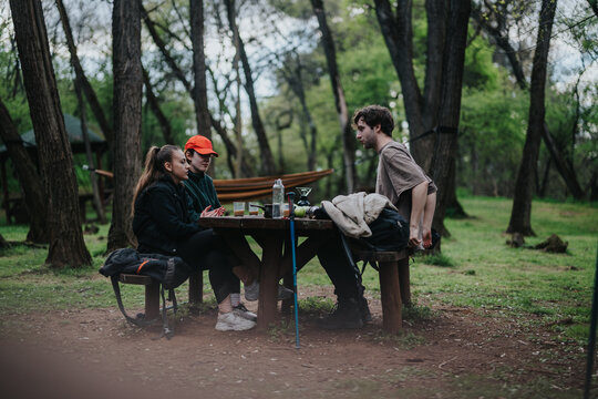 Three friends sit at a wooden picnic table in a green forest, enjoying drinks and snacks while talking during a camping trip. Hammock and gear visible in a peaceful outdoor setting.