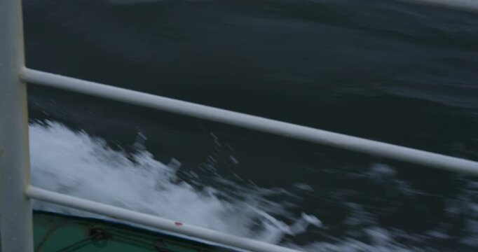Steel hull cuts through water leaving foamy trail. Waves shimmer reflecting industrial rhythm and open-sea calm. Ship railing closeup