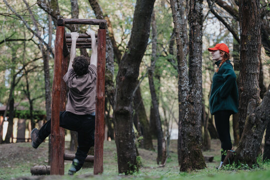 A boy climbs a rustic wooden jungle gym in a shaded park while a girl in a green coat and red cap watches nearby. An outdoor moment of fitness, play, and companionship.