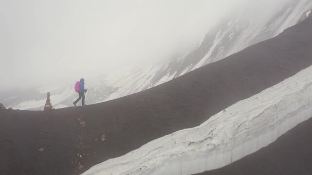 Lonely female backpacker in a rain cover with a backpack and trekking poles, making an acclimatization trek around Lenin Peak base camp and climbing Petrovsky Peak (4910 m) Pamir mountain, Kyrgystan