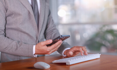 Business communication and multitasking concept. Businessman in a suit using smartphone while typing on a keyboard in a modern office, balancing digital tasks