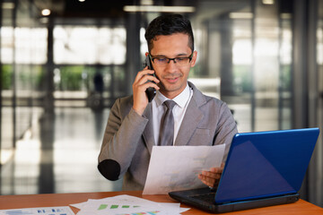 Businessman working at desk with laptop and papers, making a phone call in a modern office environment