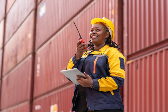 Confident female worker in safety gear uses walkie-talkie and tablet while overseeing shipping containers in logistics yard