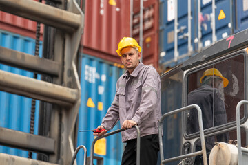 Male worker in safety gear operates heavy machinery near stacked shipping containers in an industrial cargo terminal.