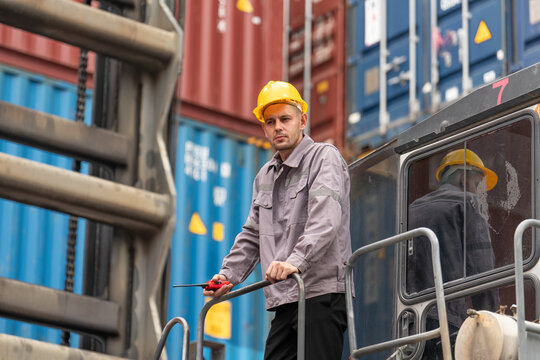 Male worker in safety gear operates heavy machinery near stacked shipping containers in an industrial cargo terminal.