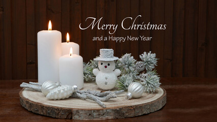 Burning white candles with Christmas baubles, a snowman and fir branches in front of a wooden board. The greeting "Merry Christmas and a Happy New Year" is written on the wood.