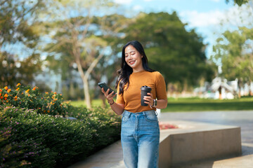 Young woman walking in park checking smartphone and holding coffee cup