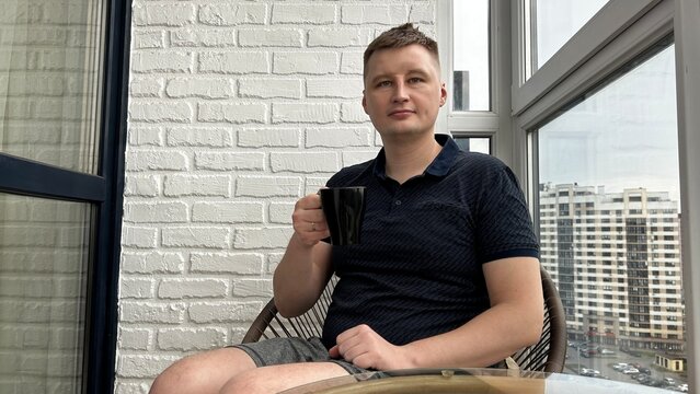 Man sitting on a balcony with a cup of coffee, enjoying a peaceful moment overlooking city buildings - Powered by Adobe
