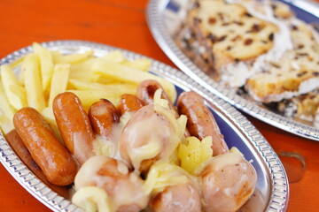 揚げたてポテトとソーセージを銀皿に盛り付けたフード写真。屋外イベントやフェス飯として親しまれる、カジュアルで楽しい食卓イメージ。温かみのある雰囲気