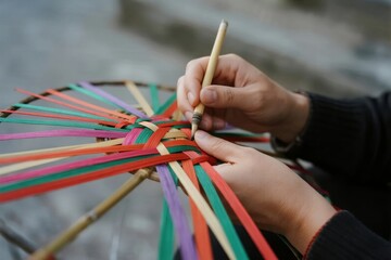 Hands weaving colorful strips of fabric into a circular pattern using a wooden tool
