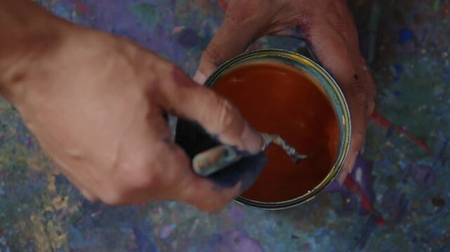 Hand r&uuml;hrt orangene Fl&uuml;ssigfarbe im Eimer um &ndash; k&uuml;nstlerischer Mischprozess mit Pigmenten, kreativer Studioumgebung und dynamischer Farbbewegung