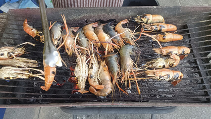 Grilled prawns on charcoal barbecue with tongs flipping seafood, smoky outdoor market scene,...