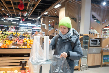 A teenage boy in winter clothes stands near fruit in a store. The child tears a plastic bag from a roll of bags in a supermarket.