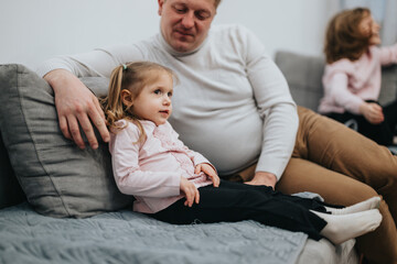 A father and his young daughter relax on a couch, enjoying quality bonding time. The warm, indoor environment highlights their connection.