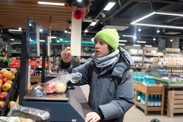 A teenage boy in winter clothes shops at a store. The child weighs items on a self-service electronic scale to check the weight, price, and to get barcode for payment.