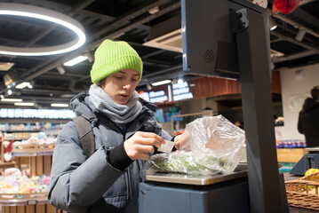 A teenage boy in winter clothes at a grocery store attaches a sticky barcode to a bag of cucumbers printed by a self-service electronic scale for payment.