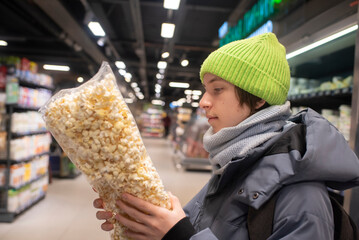 A teenage boy in winter clothes holds and looks at a package of popcorn in a supermarket