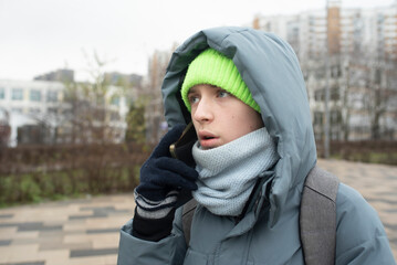 A 13-year-old teenage boy in a gray hooded jacket talks on the phone on the street in front of the school.