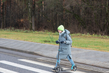 A teenage boy in a hat and jacket holds a scooter and crosses the road at a pedestrian crossing